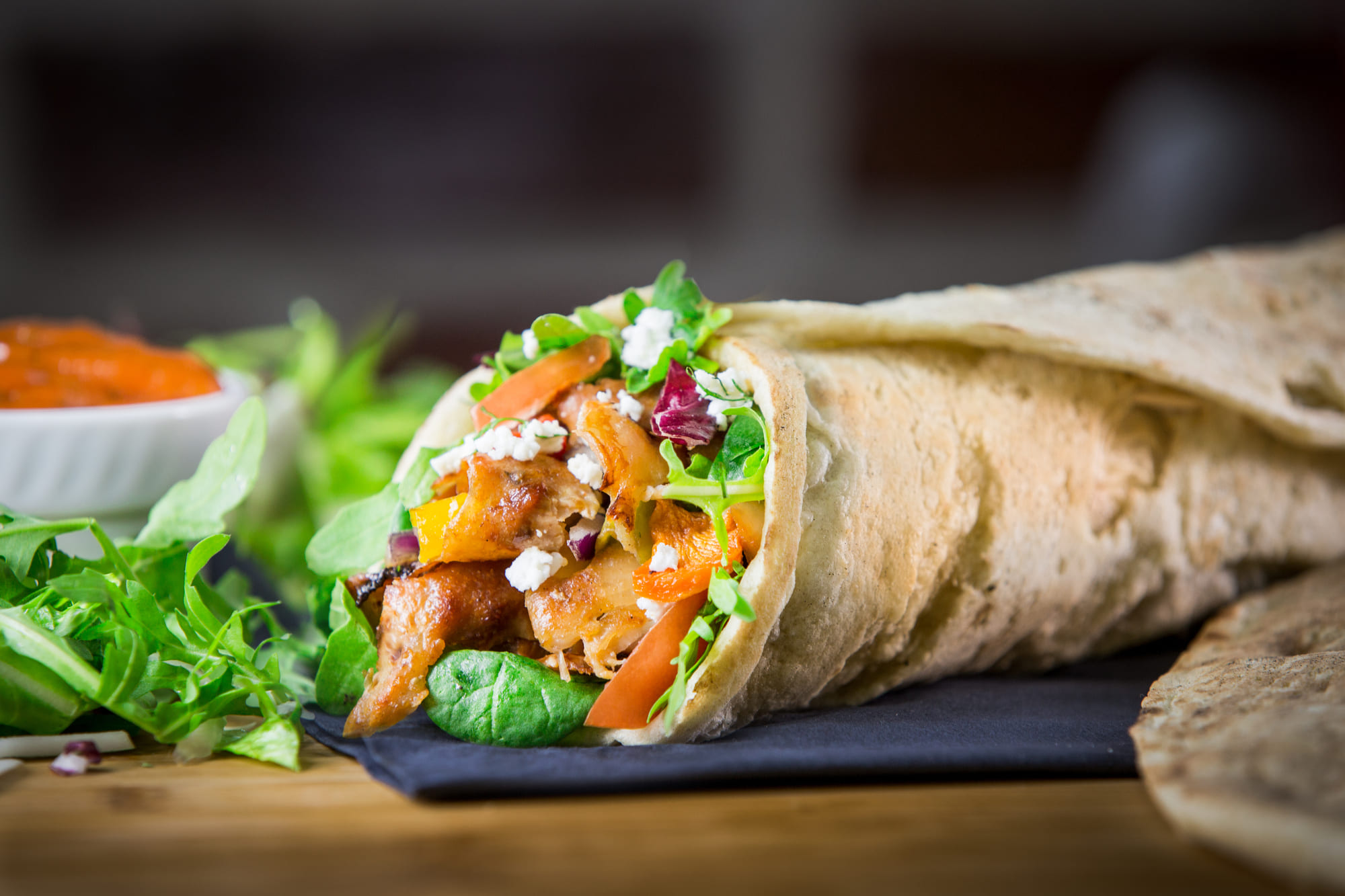 Eine Nahaufnahme eines frischen Hühnchen-Wraps mit Salat, Tomaten, Käse und gemischtem Salat auf einem Holztisch.