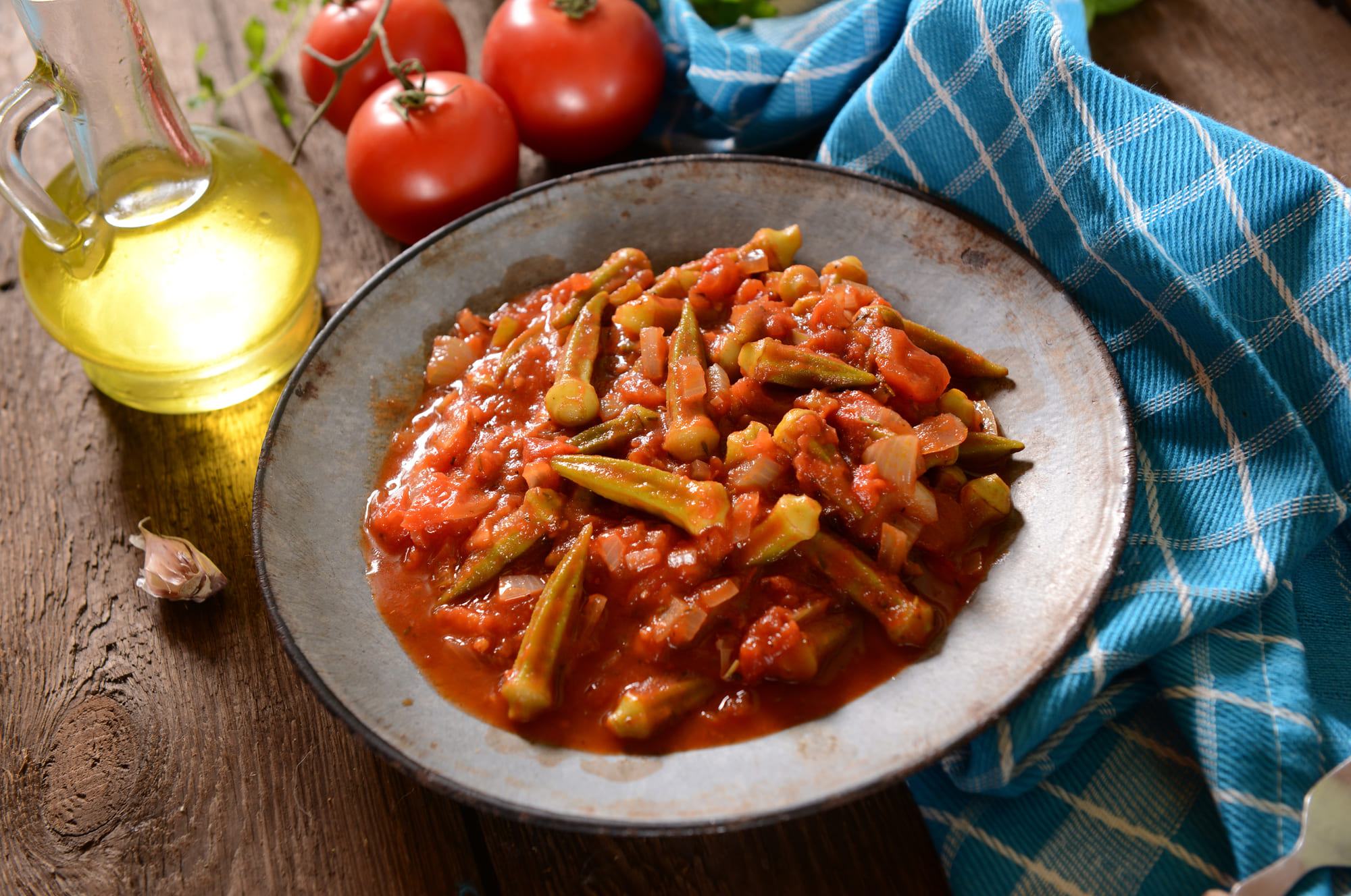 Ein Teller Okraschoteneintopf mit Tomaten, daneben frische Tomaten, Olivenöl und ein blau kariertes Tuch.