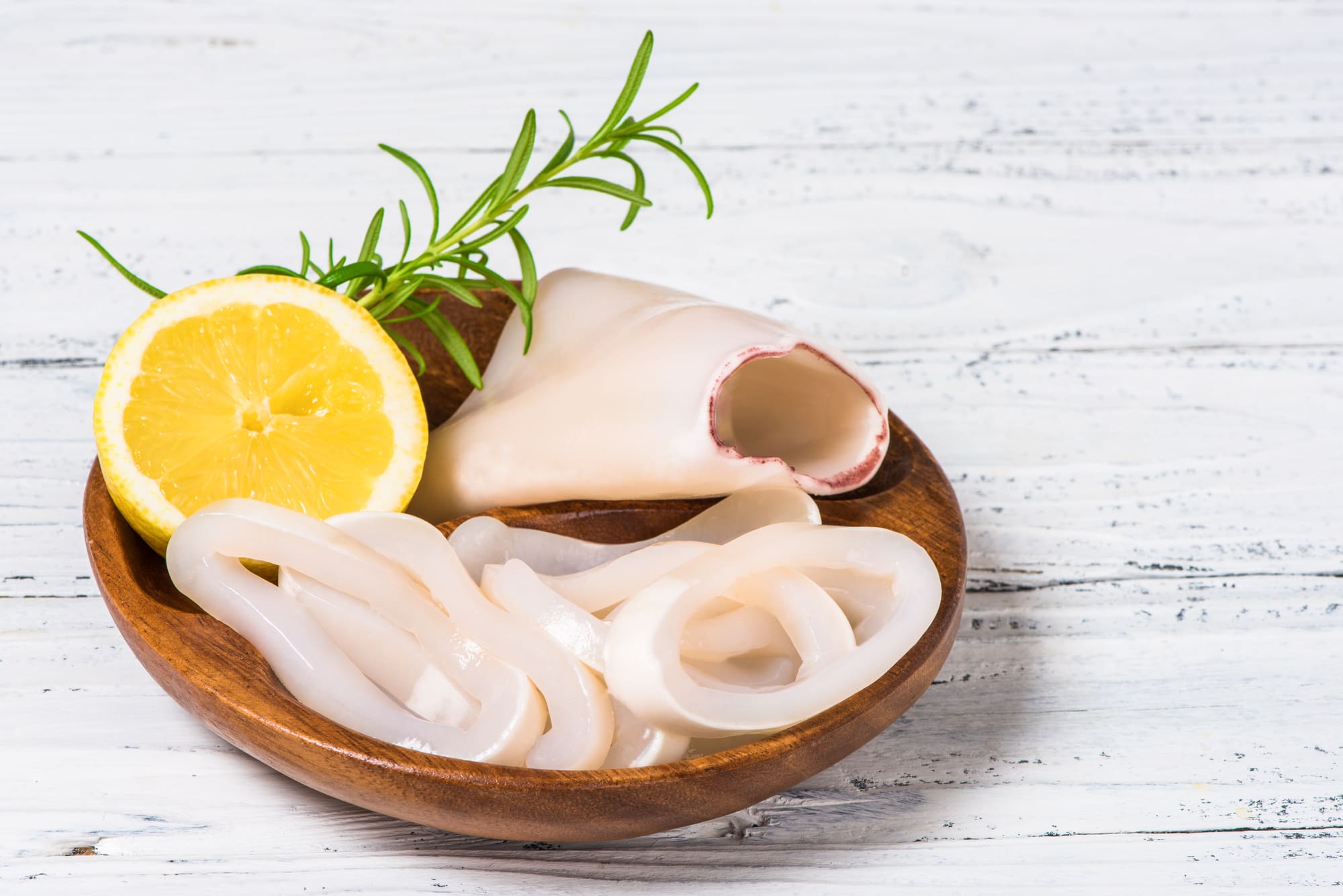 Rohe Calamari-Ringe und eine Zitronenhälfte mit Rosmarin auf einem Holzteller vor weißem, rustikalem Hintergrund.